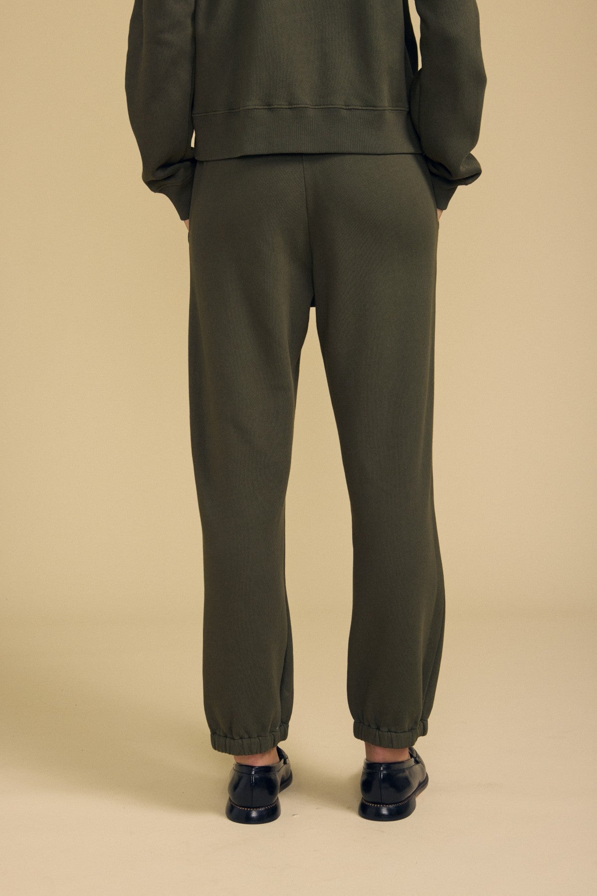 A person stands against a beige background, shown from the back, wearing the ZARA SWEATPANT in olive green by Velvet by Graham & Spencer. Featuring a relaxed fit and paired with black slip-on shoes, these sweatpants are crafted from soft cotton fleece for comfort and effortless style.-41838117814465