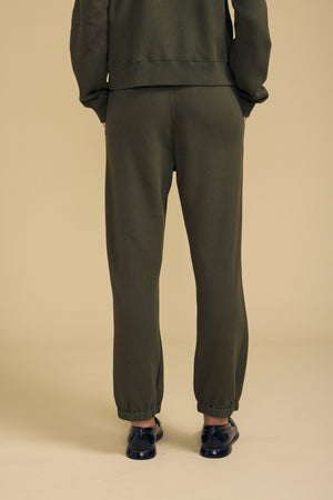 A person stands against a beige background, shown from the back, wearing the ZARA SWEATPANT in olive green by Velvet by Graham & Spencer. Featuring a relaxed fit and paired with black slip-on shoes, these sweatpants are crafted from soft cotton fleece for comfort and effortless style.