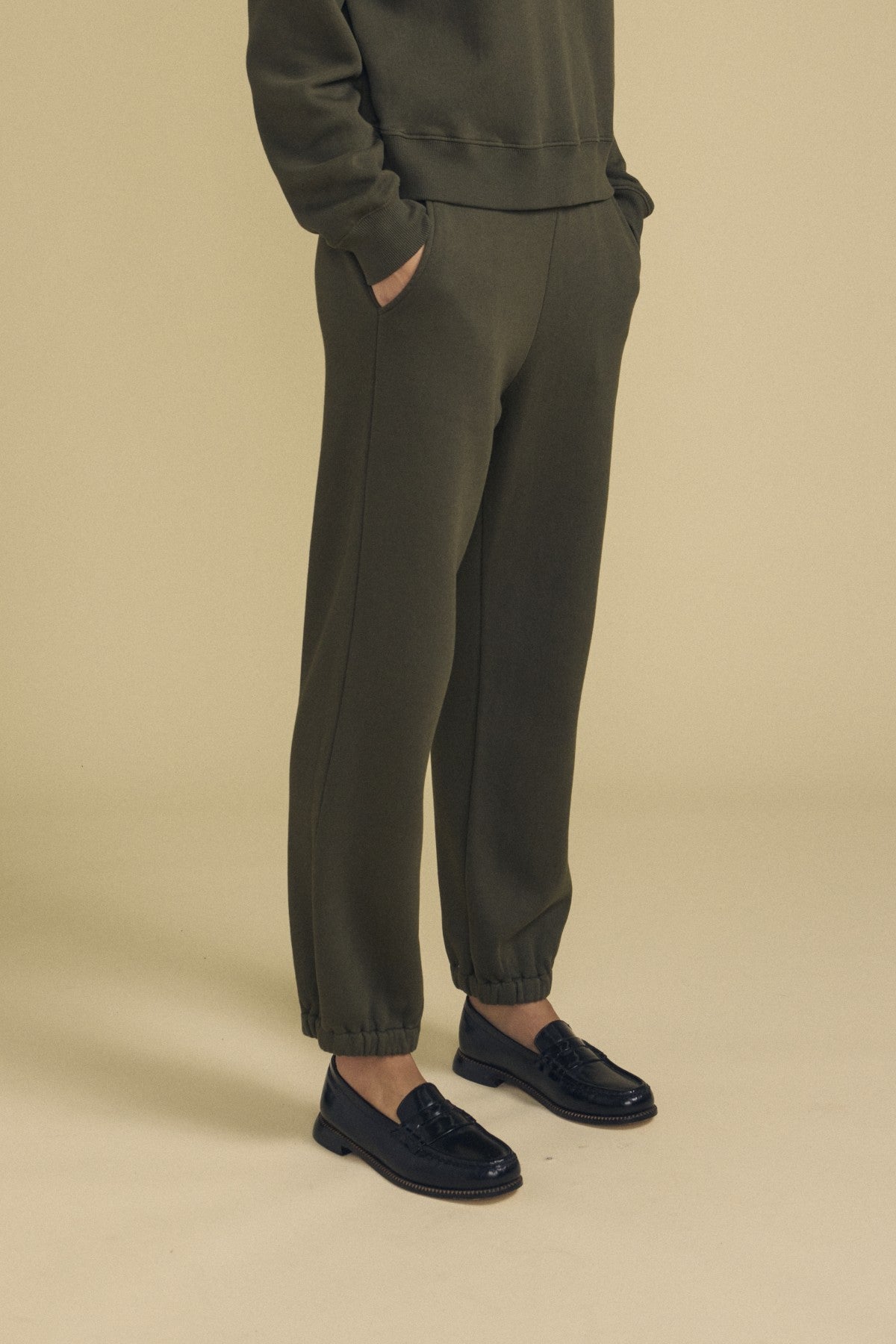 Model wearing olive green ZARA SWEATPANT by Velvet by Graham & Spencer in soft cotton fleece, paired with a matching sweatshirt and black dress shoes, standing with hands in pockets against a beige background for a relaxed fit look.