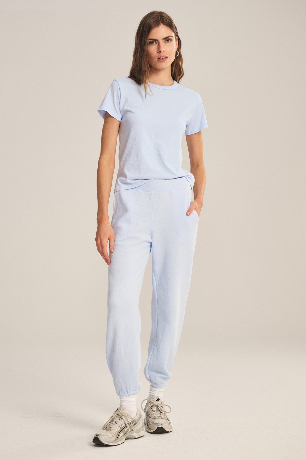 A woman stands in a relaxed pose against a plain beige background, wearing a light blue T-shirt, ZARA SWEATPANT by Velvet by Graham & Spencer—relaxed fit sweatpants crafted from soft cotton fleece—and white sneakers.
