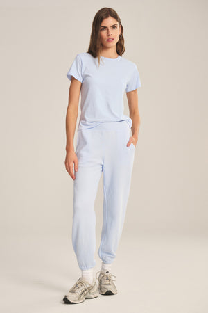 A woman stands in a relaxed pose against a plain beige background, wearing a light blue T-shirt, ZARA SWEATPANT by Velvet by Graham & Spencer—relaxed fit sweatpants crafted from soft cotton fleece—and white sneakers.