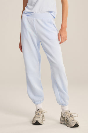 A person wears Velvet by Graham & Spencer ZARA SWEATPANT joggers in light blue cotton fleece, styled with white socks and gray sneakers, standing against a plain beige background.