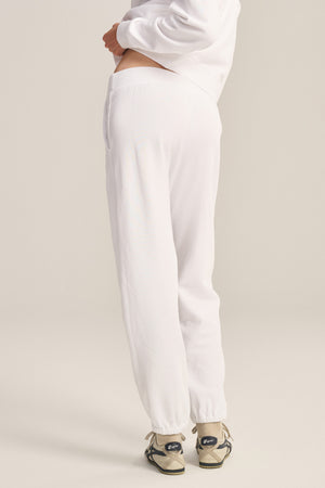 A person stands at an angle on a light background, wearing Velvet by Graham & Spencer ZARA SWEATPANT in relaxed white cotton fleece, paired with a matching sweatshirt and beige sneakers for a comfortable, effortless look.