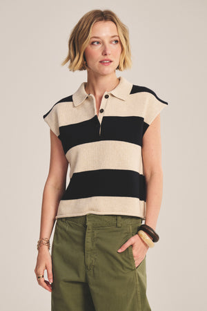 A person wears the ZOELLA SWEATER POLO by Velvet by Graham & Spencer, featuring wide black and beige stripes, paired with olive green pants and bracelets, standing against a plain background.