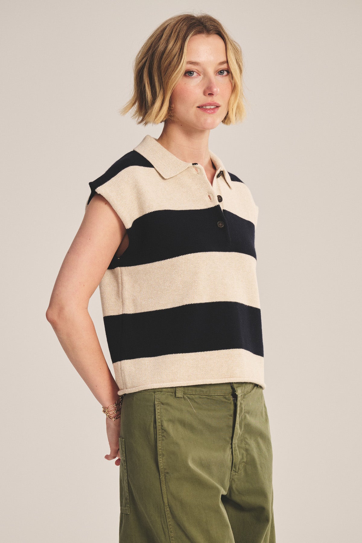 A woman with short blonde hair wears the ZOELLA SWEATER POLO by Velvet by Graham & Spencer, featuring wide black and beige stripes, paired with olive green pants against a plain background.