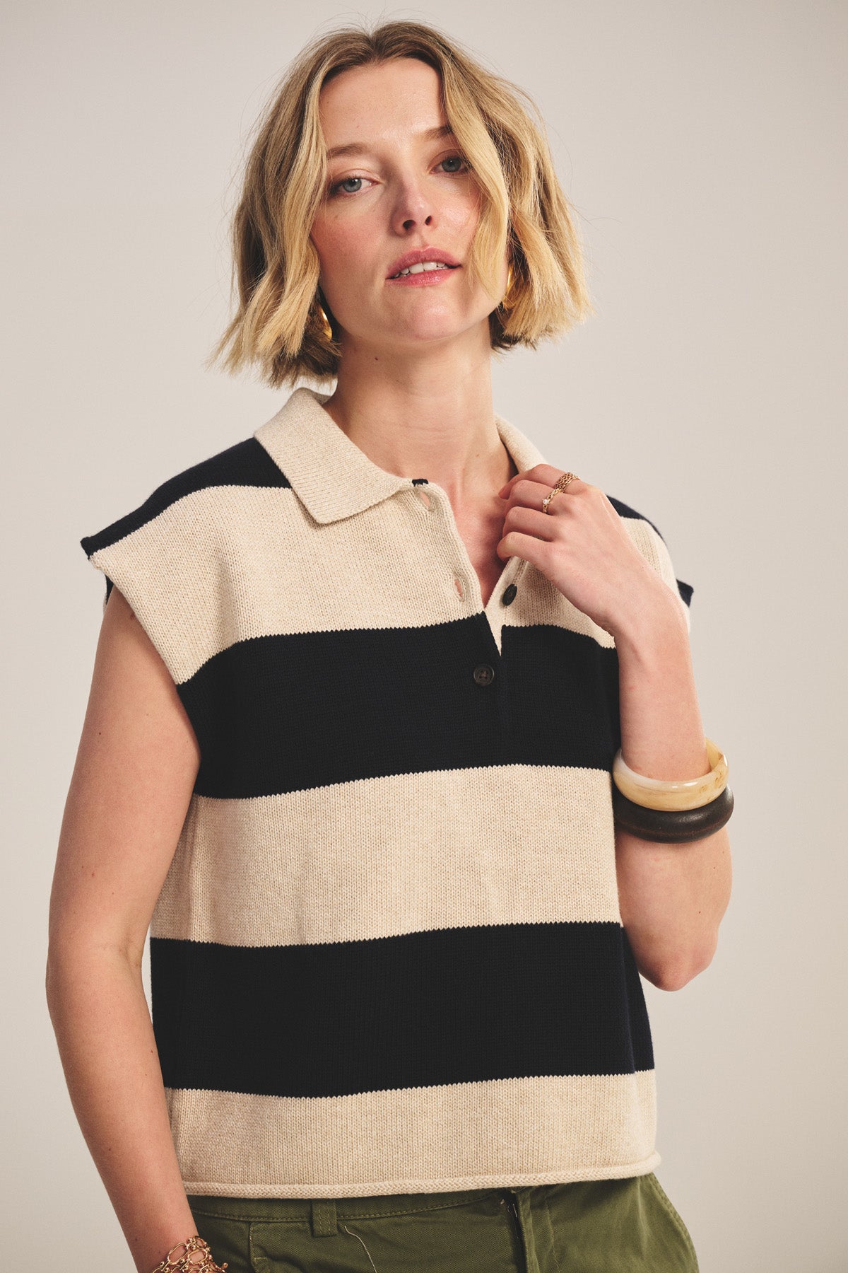 A woman with short blonde hair wears the Velvet by Graham & Spencer ZOELLA SWEATER POLO—a sleeveless, collared, black and beige striped cropped top—paired with green pants and jewelry, standing against a plain background.-40910161510593