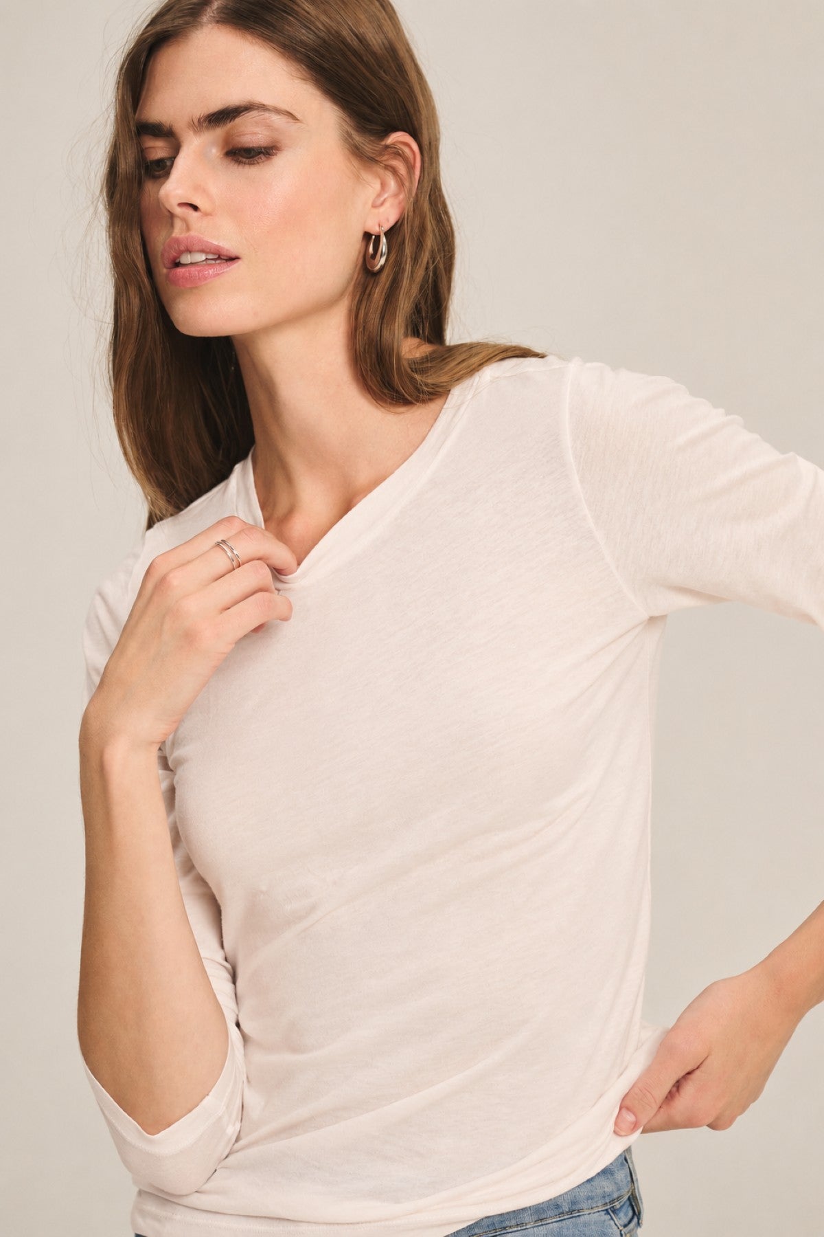 A woman with long brown hair wears the ZOFINA TEE by Velvet by Graham & Spencer, an ultra-soft gauzy whisper fitted long sleeve top with a classic crew neckline, paired with blue jeans. She stands with one hand near her collar and the other on her hip, looking slightly to the side.-41781415018689