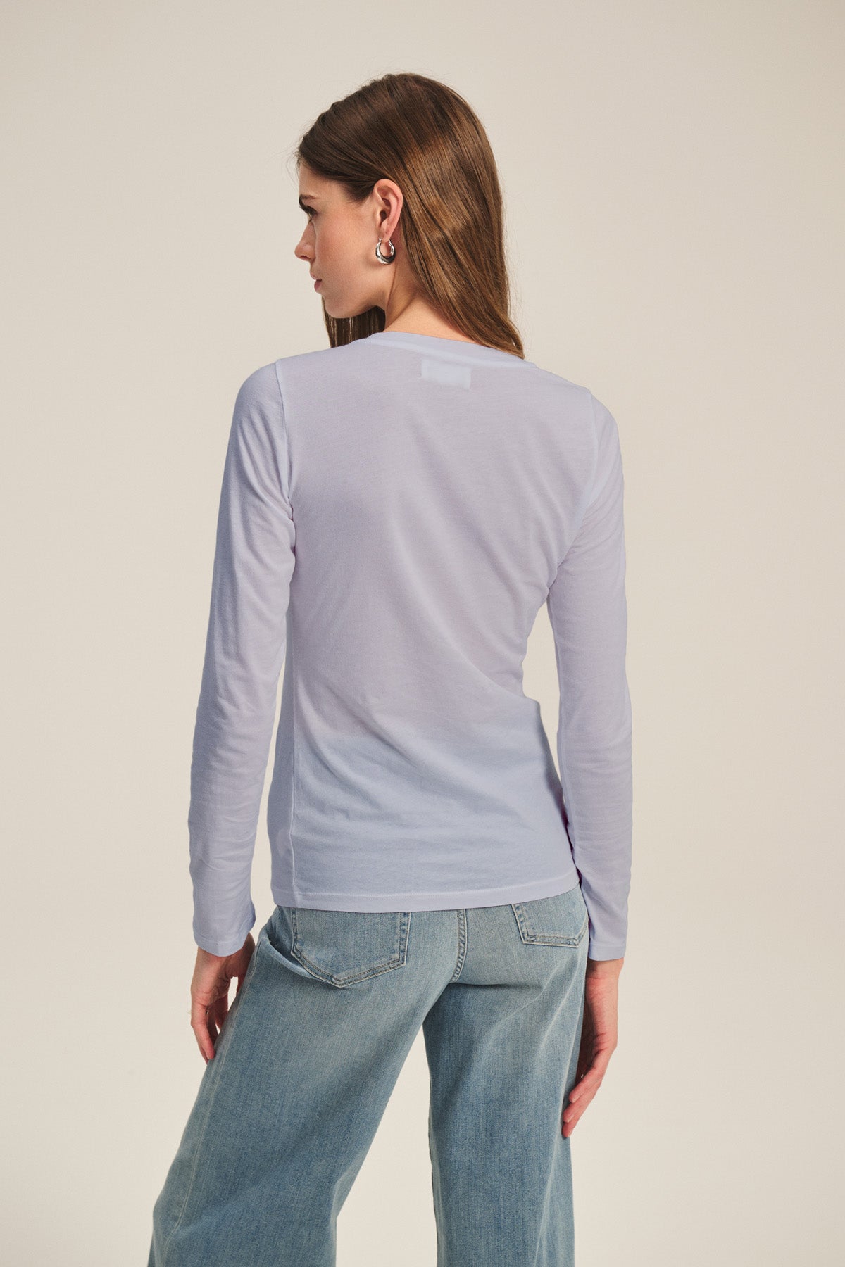 A woman with long brown hair stands with her back to the camera wearing the ZOFINA TEE by Velvet by Graham & Spencer—an ultra-soft gauzy whisper fitted long sleeve tee with a crew neckline—paired with blue jeans against a plain background.-40807729692865