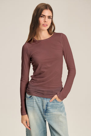 A person in the ZOFINA TEE by Velvet by Graham & Spencer, an ultra-soft gauzy whisper mauve fitted long sleeve top, paired with light blue jeans, stands against a plain background facing the camera with one hand in their pocket.