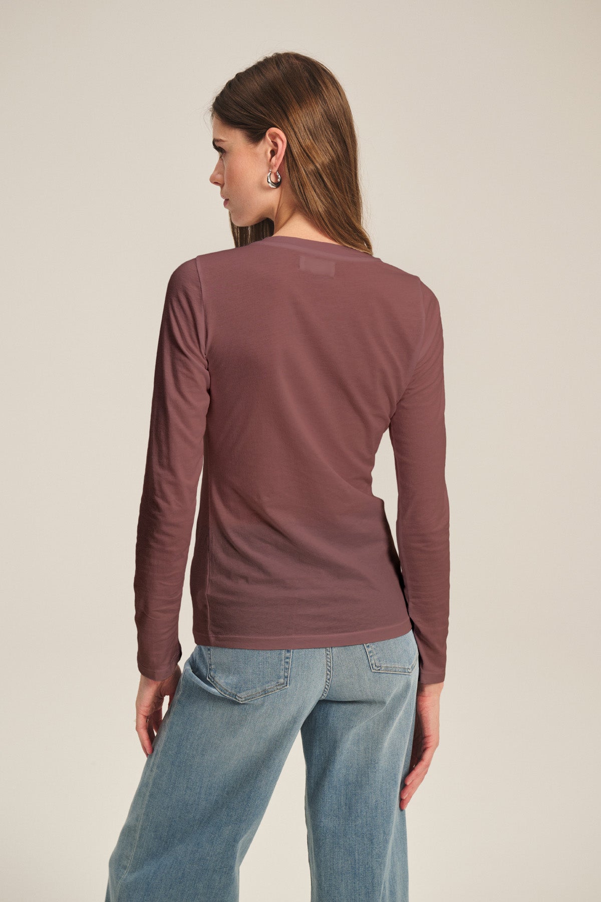 A woman faces away wearing the ZOFINA TEE by Velvet by Graham & Spencer, a fitted mauve long sleeve crew neck top, paired with light blue jeans against a neutral background.