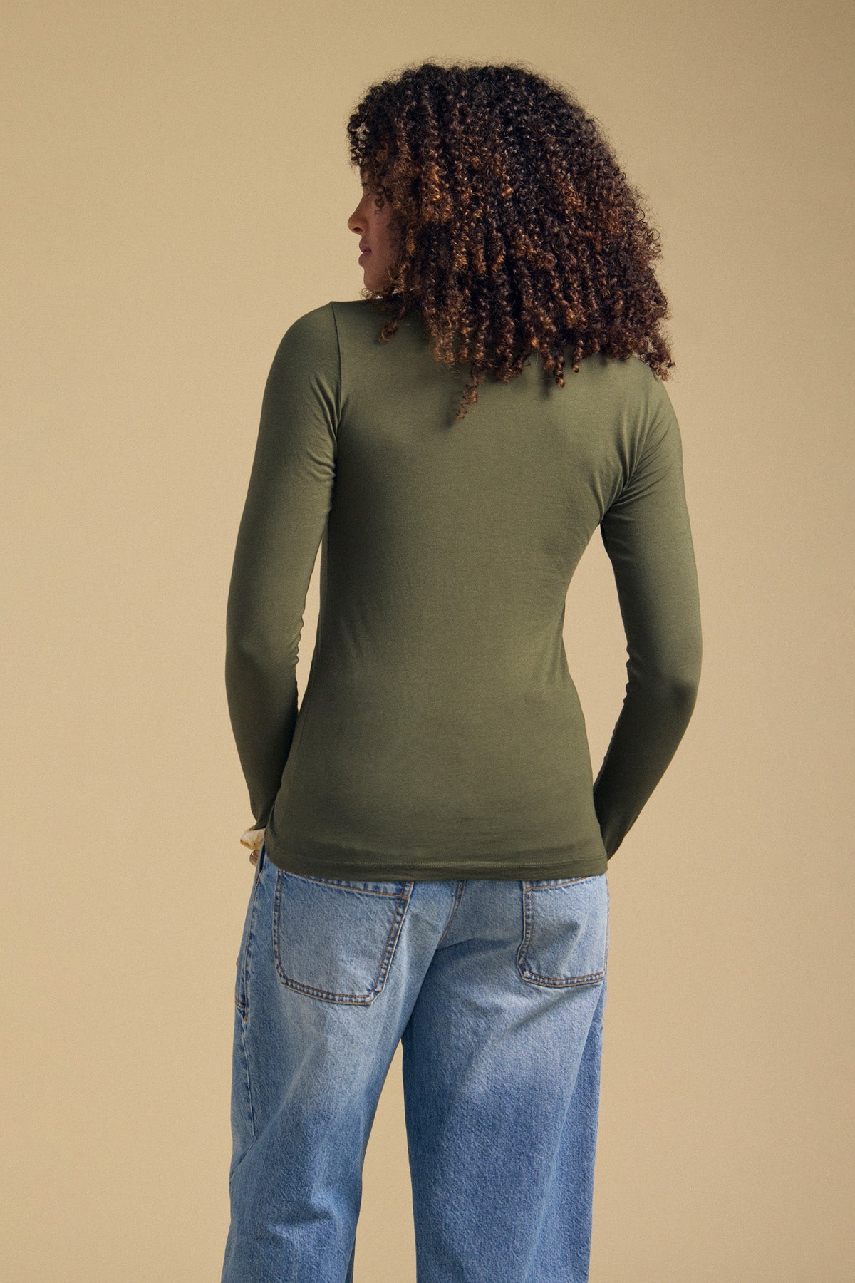 A person stands with their back to the camera, wearing the ZOFINA TEE by Velvet by Graham & Spencer—an olive green fitted long sleeve top featuring a classic crew neckline—paired with light blue jeans, set against a plain tan background.