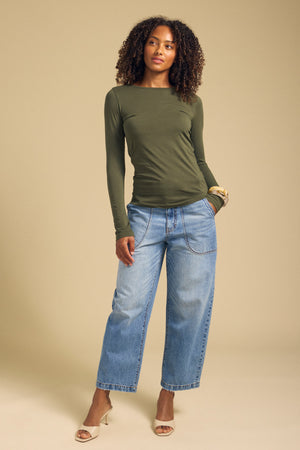 A person stands against a beige background wearing the Velvet by Graham & Spencer ZOFINA TEE—an ultra-soft, gauzy whisper olive fitted long sleeve top with a classic crew neckline—paired with wide-leg blue jeans and beige heeled sandals, with one hand in their pocket.