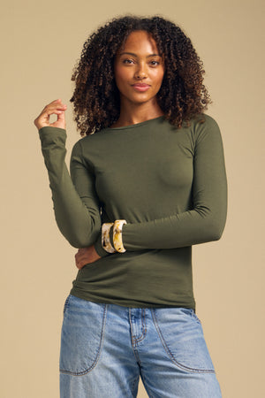 A woman poses against a beige background, resting one hand near her face, wearing the ZOFINA TEE by Velvet by Graham & Spencer—an olive green fitted long sleeve shirt with a classic crew neckline—paired with blue jeans.