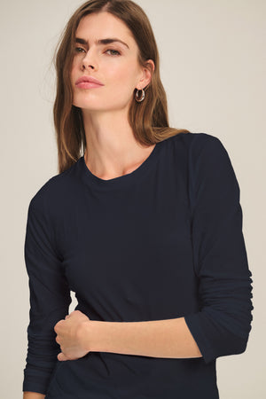 A woman with long brown hair models the ZOFINA TEE by Velvet by Graham & Spencer, an ultra-soft gauzy whisper fitted long sleeve crewneck, standing against a plain light background.