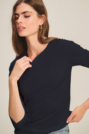 A woman wears the ZOFINA TEE by Velvet by Graham & Spencer—a fitted, navy long sleeve crewneck—paired with light jeans, standing against a neutral background with her hand near her face.