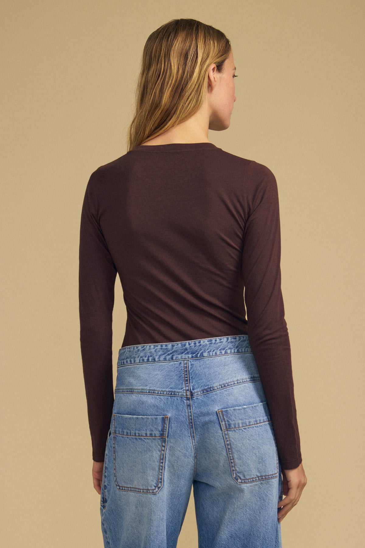 A person with long hair is seen from behind, standing against a beige background, wearing the Velvet by Graham & Spencer ZOFINA TEE—an ultra-soft gauzy whisper brown fitted long sleeve tee with a classic crew neckline—paired with blue jeans.-42063865610433