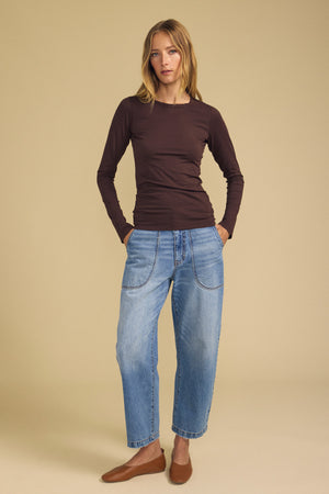A woman stands with her hands in her pockets, wearing the ZOFINA TEE by Velvet by Graham & Spencer—an ultra-soft, gauzy whisper long-sleeve brown shirt with a classic crew neckline—paired with loose blue jeans and brown shoes against a plain beige background.