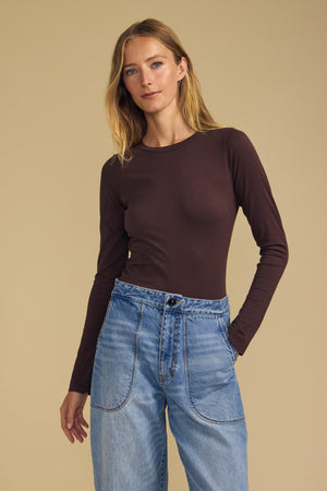 Wearing the ZOFINA TEE by Velvet by Graham & Spencer—a classic crew neckline, fitted long sleeve tee in rich brown—a woman stands against a beige background with one hand in the pocket of her high-waisted blue jeans.