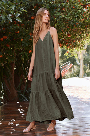 A woman in the CLEO LINEN MAXI DRESS by Velvet by Graham & Spencer stands on a wooden deck, surrounded by orange trees. She wears sandals and looks to the side.