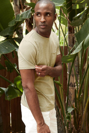 Side view of Chad Raw Edge Cotton Slub Pocket Tee in Olivine with white denim model standing outside near plants