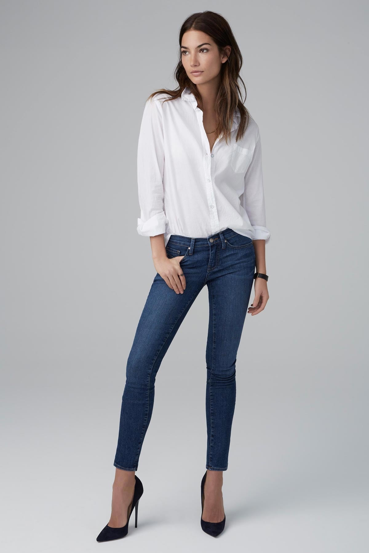 A woman stands against a plain background wearing a white button-up shirt, Velvet by Graham & Spencer's dark blue TONI SKINNY JEAN, and black high-heeled shoes. She has one hand in her pocket and is looking slightly to the side, comfy and looking good.-1176066031697