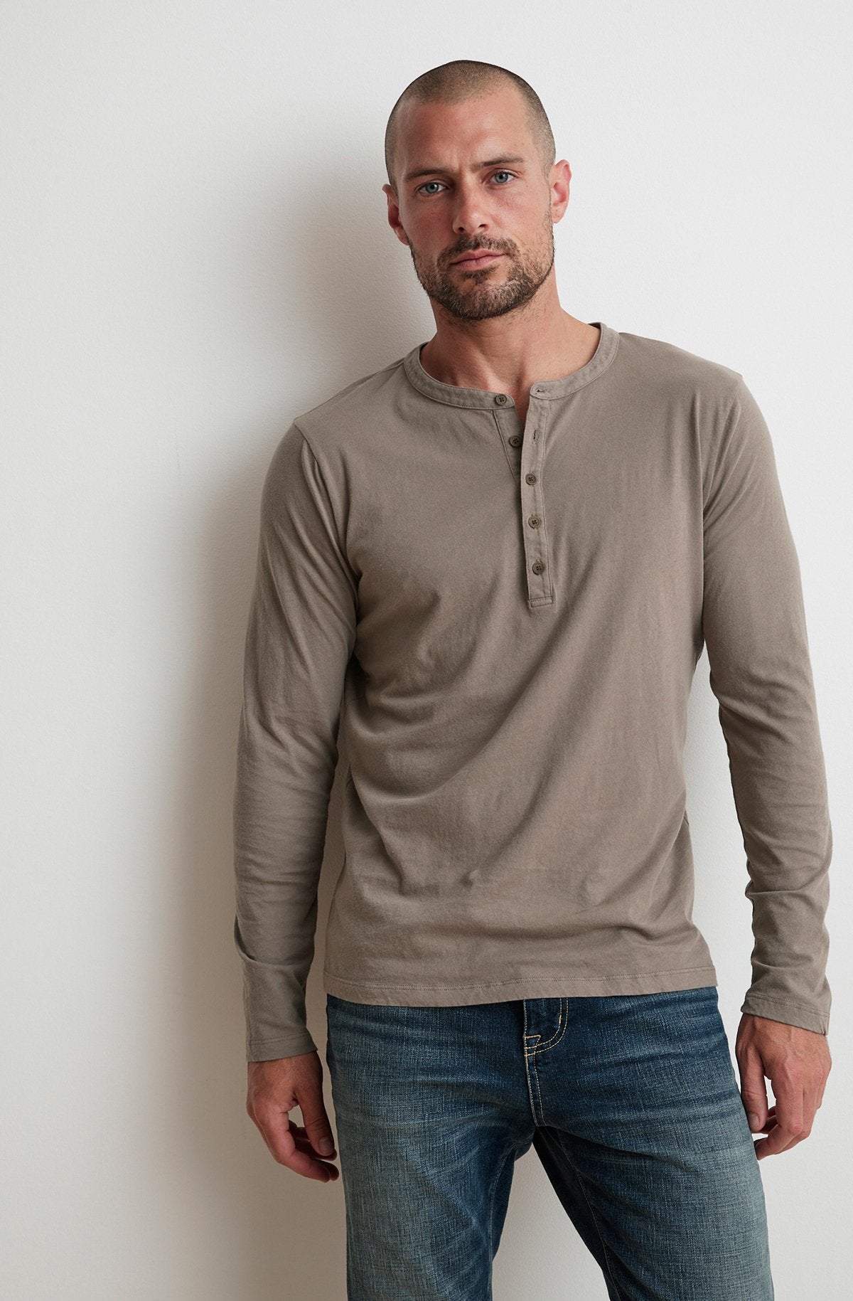 A man in an ALVARO HENLEY by Velvet by Graham & Spencer and blue jeans stands against a white wall.-23687315914945