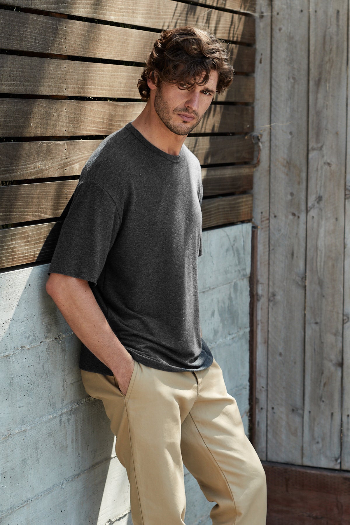   Model leaning against a wall outside with hands in pockets wearing Newall crew neck shirt in dark heathered grey anthracite color. 