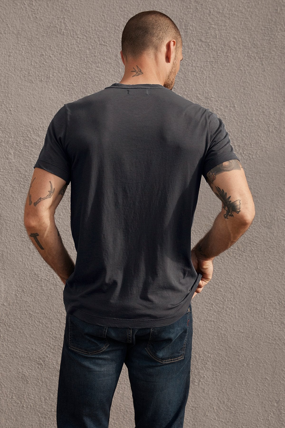 A person with short hair, wearing a Velvet by Graham & Spencer SAMSEN WHISPER CLASSIC V-NECK TEE and jeans—perfect for everyday wear—is standing with their back to the camera, showcasing various arm tattoos.-23695162540225