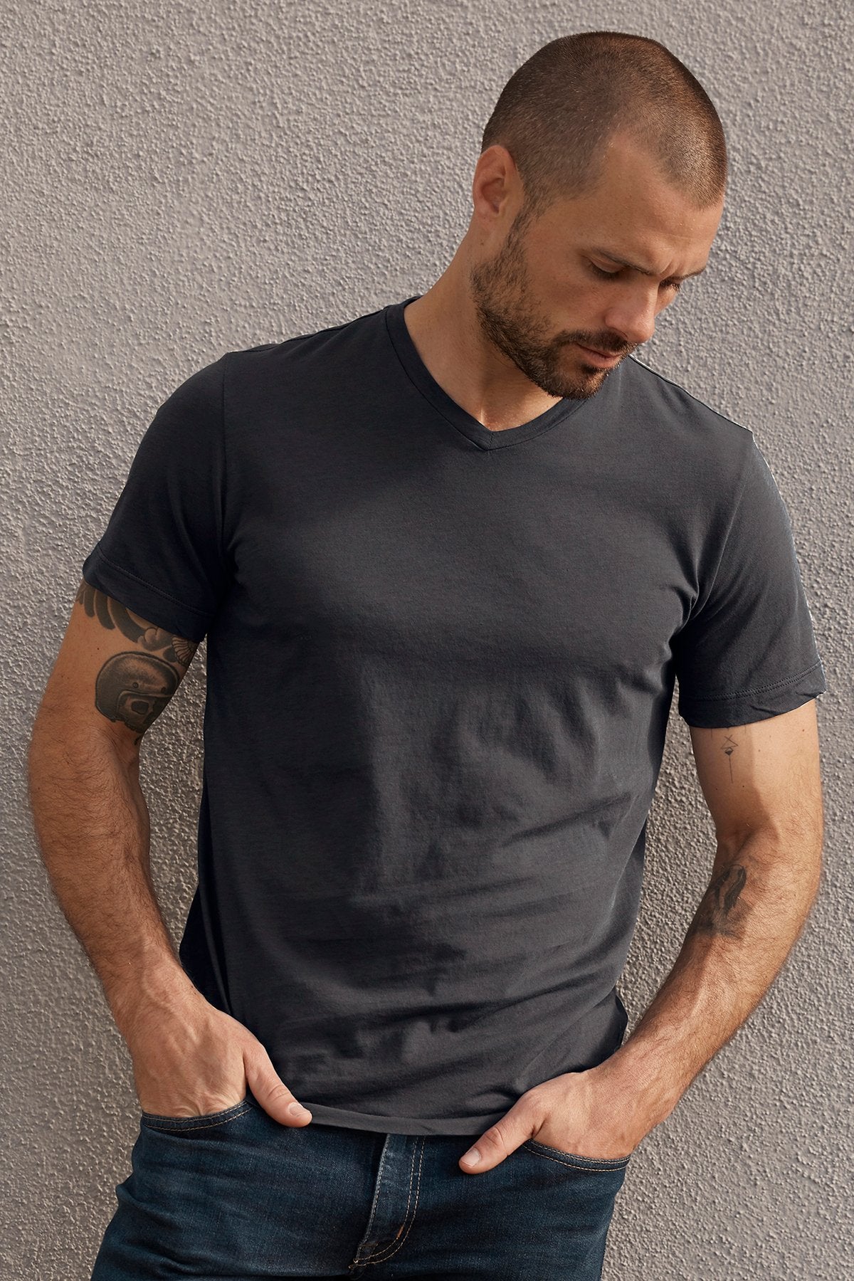   A man with short hair and tattoos on his arms wears a dark gray SAMSEN WHISPER CLASSIC V-NECK TEE by Velvet by Graham & Spencer and jeans, standing against a textured gray wall. 