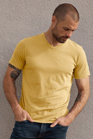 A man with a shaved head and tattoos on his arms is wearing a yellow SAMSEN WHISPER CLASSIC V-NECK TEE by Velvet by Graham & Spencer and dark jeans. He stands against a textured wall, looking down.