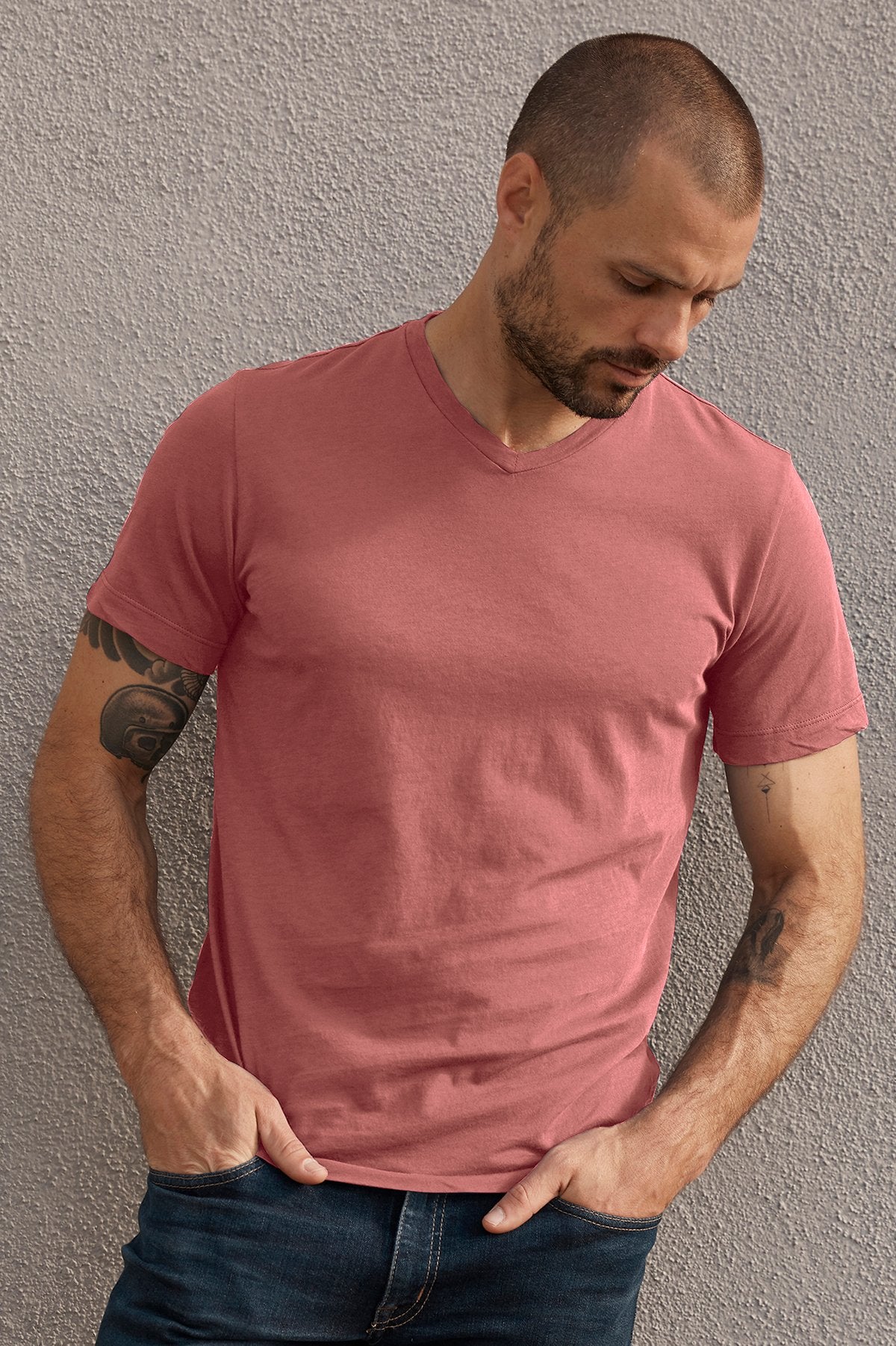   A man with a shaved head, beard, and tattoos on his left arm is wearing a pink SAMSEN WHISPER CLASSIC V-NECK TEE by Velvet by Graham & Spencer and jeans while standing against a textured gray wall. 