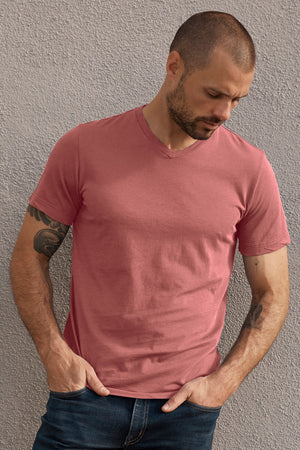 A man with a shaved head, beard, and tattoos on his left arm is wearing a pink SAMSEN WHISPER CLASSIC V-NECK TEE by Velvet by Graham & Spencer and jeans while standing against a textured gray wall.