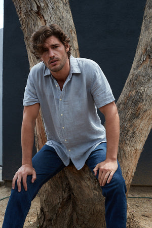 A man in a Velvet by Graham & Spencer MACKIE LINEN BUTTON-UP SHIRT leaning against a tree.