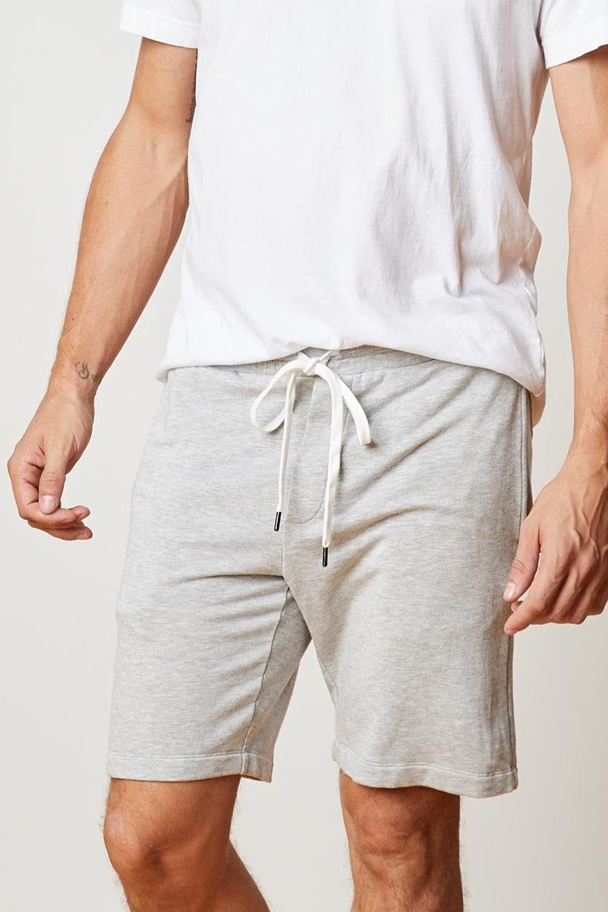 A person wearing a white t-shirt and light gray ATLAS LUXE FLEECE DRAWSTRING SHORT by Velvet by Graham & Spencer, featuring tailored design details, is standing with their arms slightly bent. The background is plain beige.-25052221145281