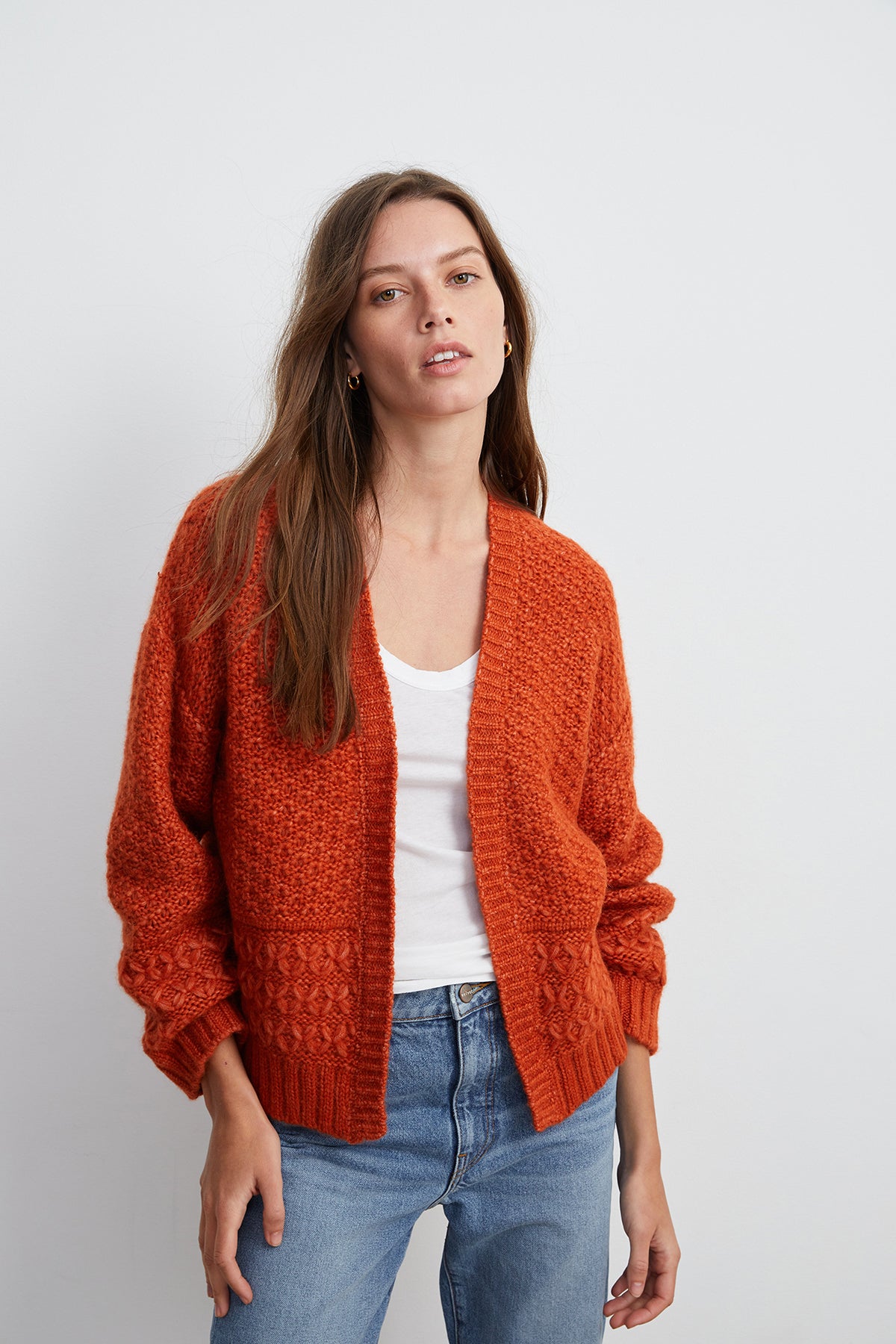   A woman wearing the Velvet by Graham & Spencer YUNA OPEN CARDIGAN, an orange open-knit piece made from a cozy alpaca and wool blend, poses against a plain white background. She has long brown hair and is also wearing a white shirt and blue jeans, creating a relaxed and handstitched feel. 