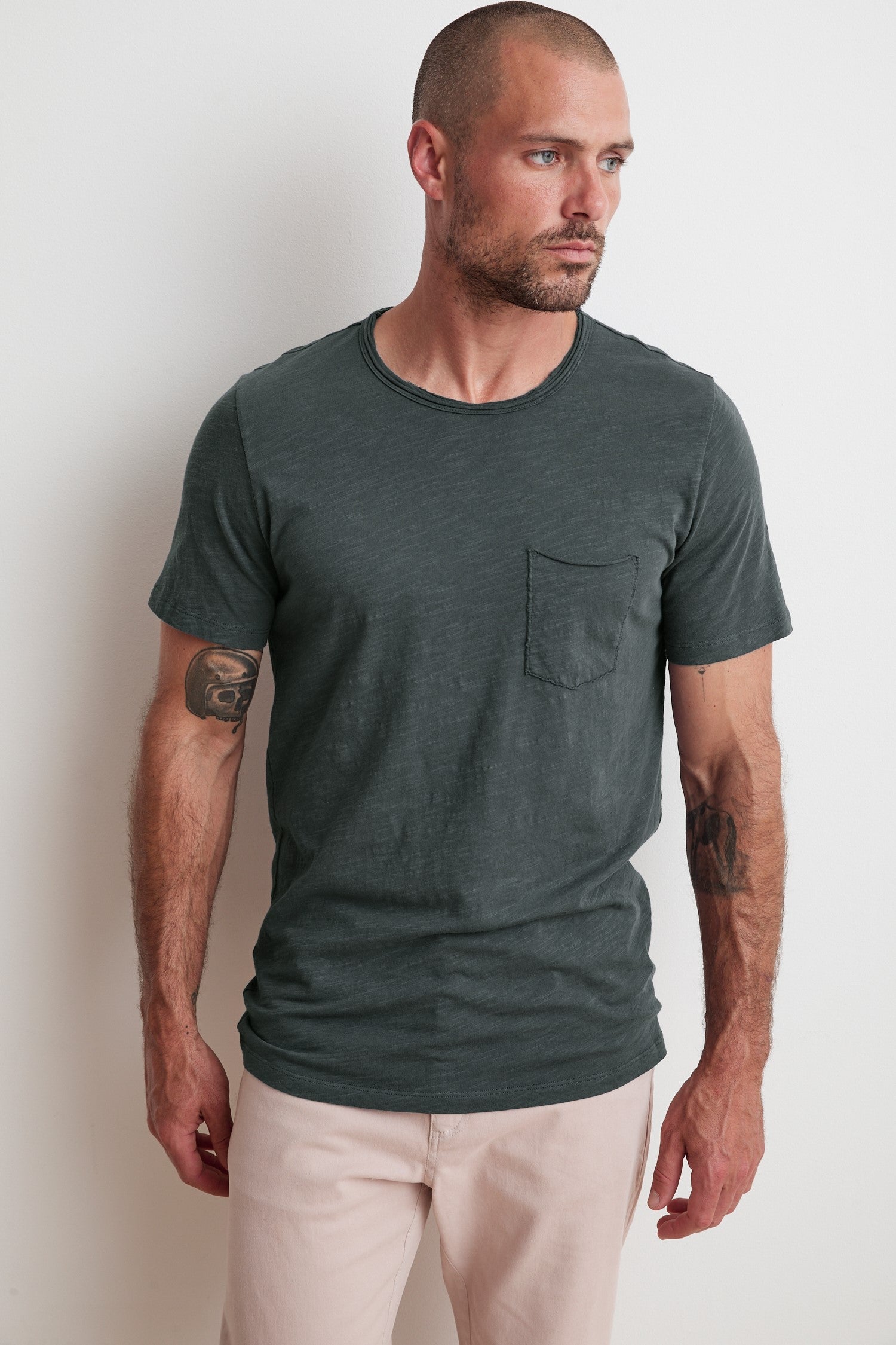   A man with short hair and a beard wearing a dark green CHAD TEE from Velvet by Graham & Spencer made from textured cotton slub and light-colored pants stands against a plain white wall, looking to his right. He has tattoos on his right arm. 