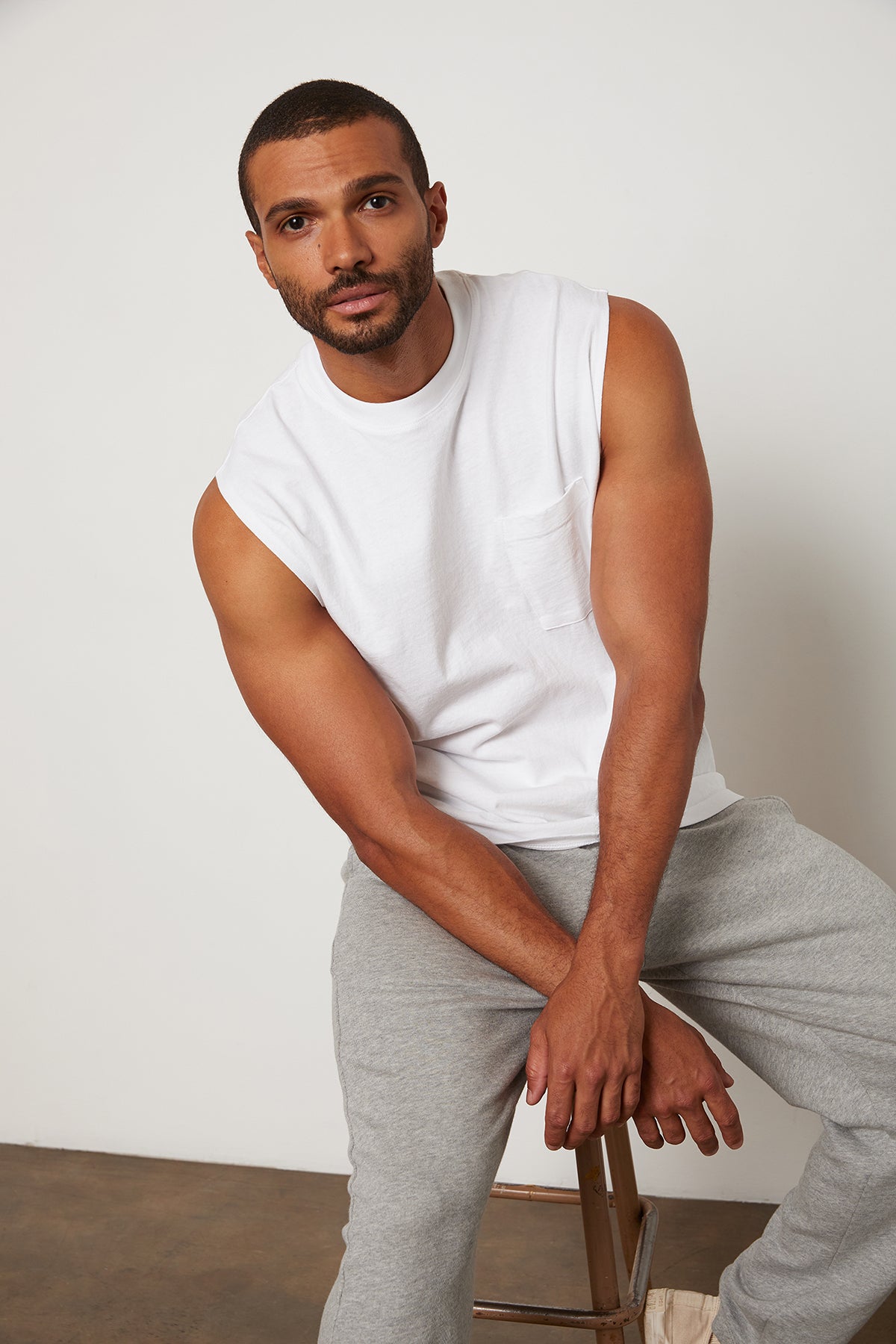 Bodhi Crew Neck Muscle Tee in White with Judas Pant in Heather Grey with Model Sitting on Stool.-24705813250241
