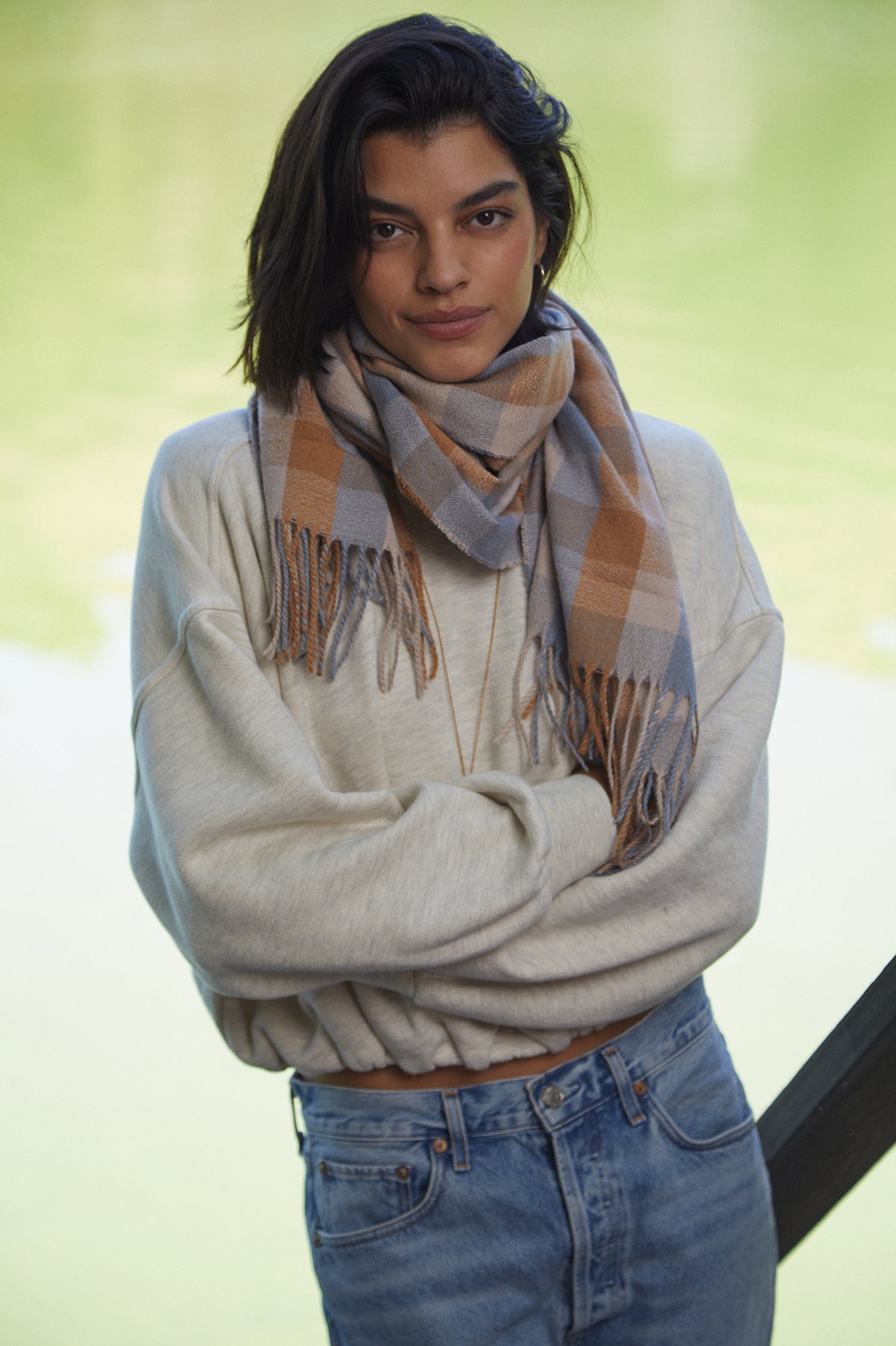   A person stands with arms crossed, wearing a light-colored sweater, a CHECK SCARF by Velvet by Graham & Spencer, and blue jeans, against a backdrop of greenery and water. 