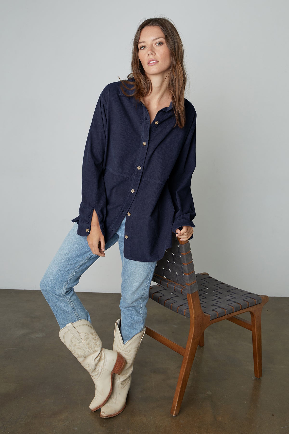   Model leaning on chair wearing Jeslyn Corduroy Button-Up Shirt in midnight blue with light blue jeans and cream colored cowboy boots. 