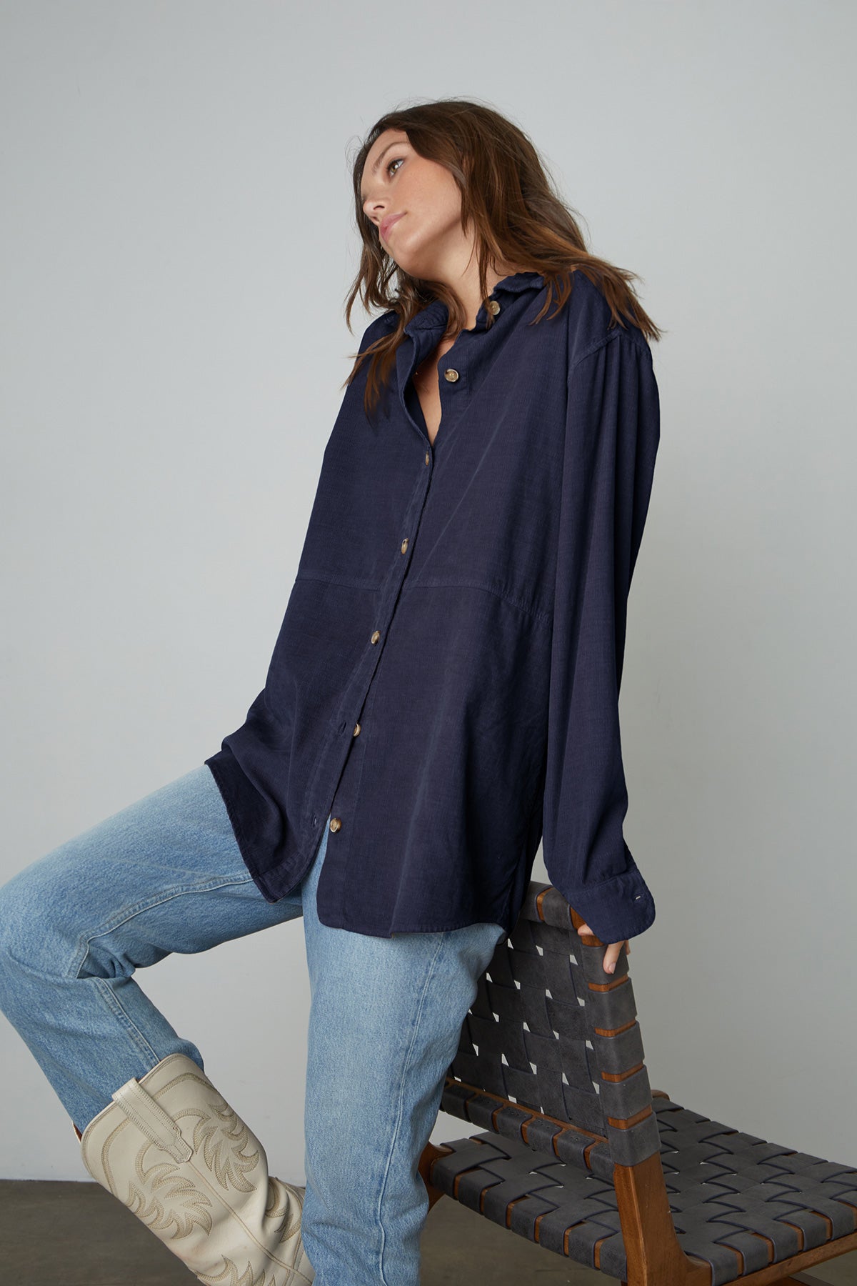   Model leaning on chair wearing Jeslyn Corduroy Button-Up Shirt in midnight blue with light blue jeans and cream colored cowboy boots side and front 