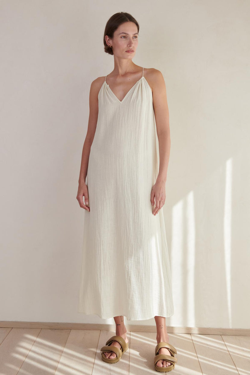 A woman in a long, white, sleeveless Carrillo dress stands in a room with soft lighting, wearing golden sandals by Velvet by Jenny Graham.
