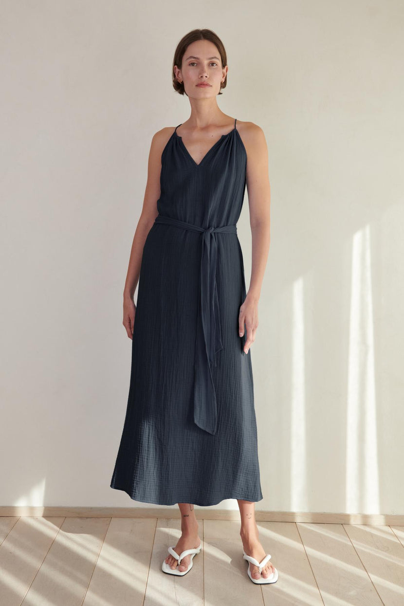A woman in a navy blue sleeveless Carrillo dress by Velvet by Jenny Graham and white slides, standing in a softly lit room with shadows on the wall.