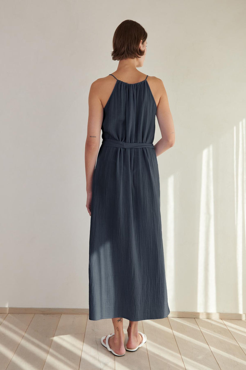 A woman stands facing away from the camera in a sunlit room, wearing a navy maxi Carrillo dress by Velvet by Jenny Graham with a halter neck and white sandals.