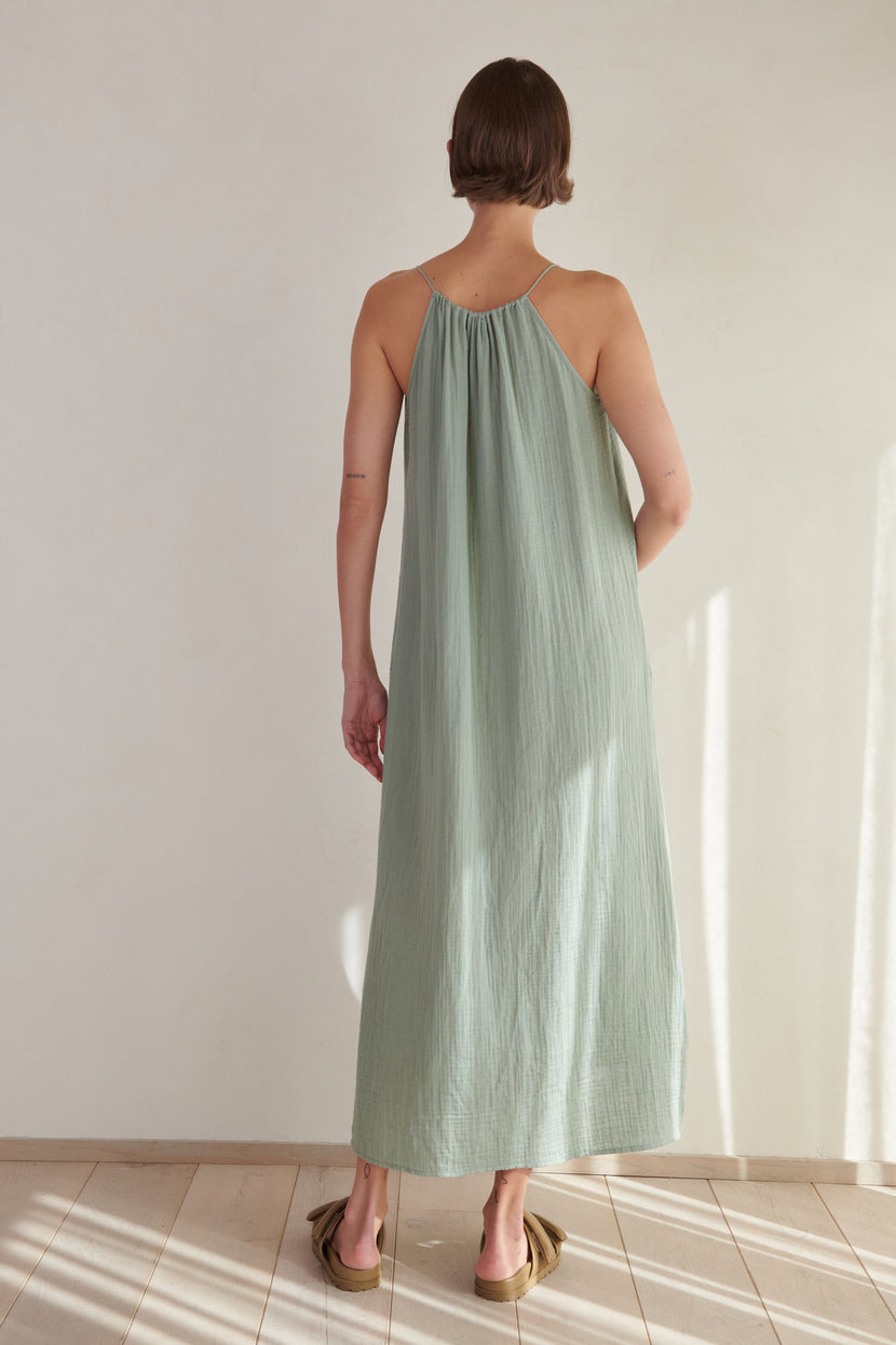 Woman standing in a sunlit room, wearing a long, light green cotton gauze Carrillo dress by Velvet by Jenny Graham with thin straps, facing away from the camera.