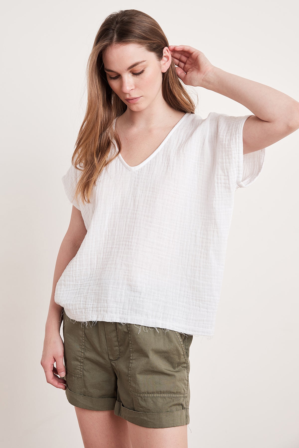 A person wearing the Velvet by Graham & Spencer ELSA Cotton Gauze Top in white and olive green shorts, looking down with one hand touching their hair. The relaxed fit and raw hem detail of the top add a casual yet stylish touch to the outfit.-7367247528017