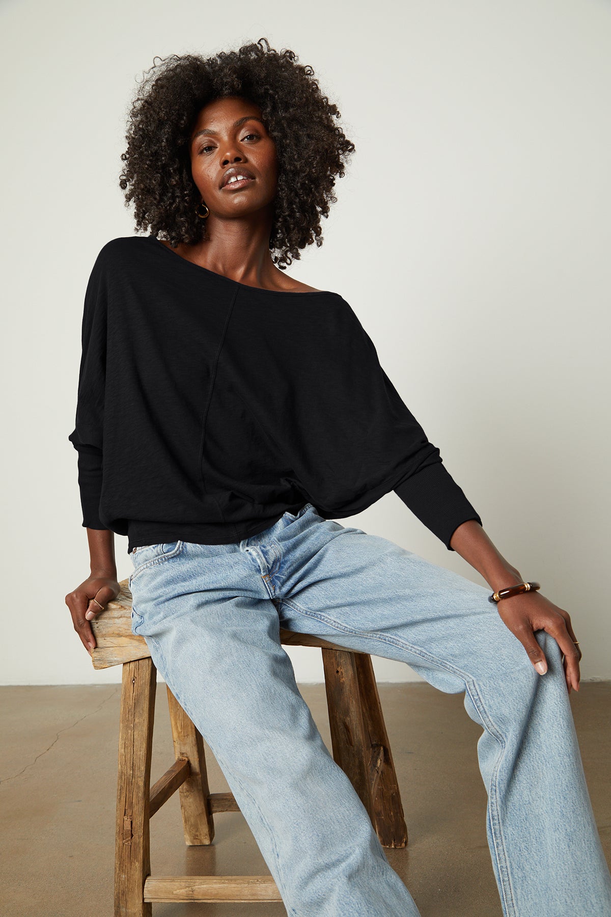 A woman is sitting on a stool wearing JOSS DOLMAN SLEEVE TEE by Velvet by Graham & Spencer jeans and a black top.-26235758313665