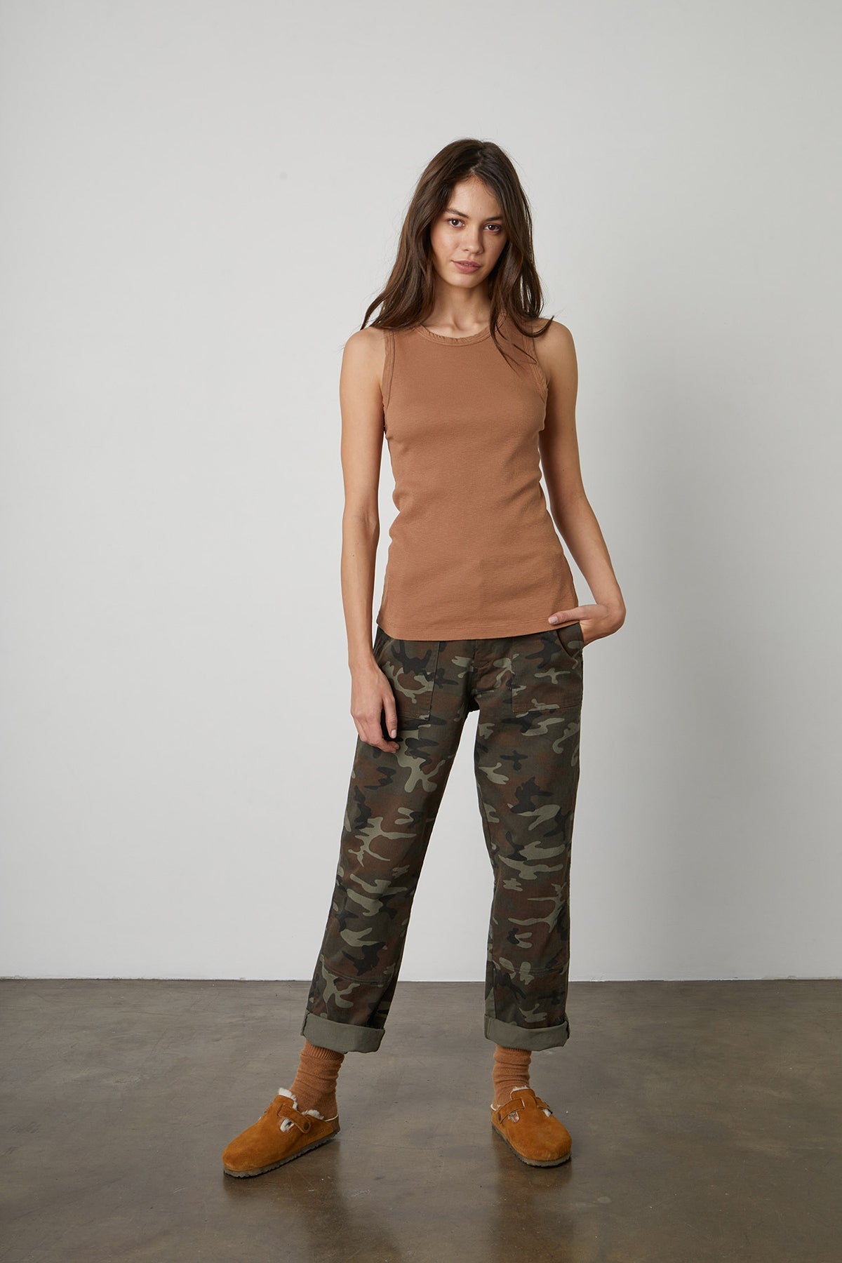   A woman stands against a plain background, wearing a brown MAXIE RIBBED TANK TOP by Velvet by Graham & Spencer, camouflage pants, matching brown socks, and tan loafers. One of her hands is in her pocket. 