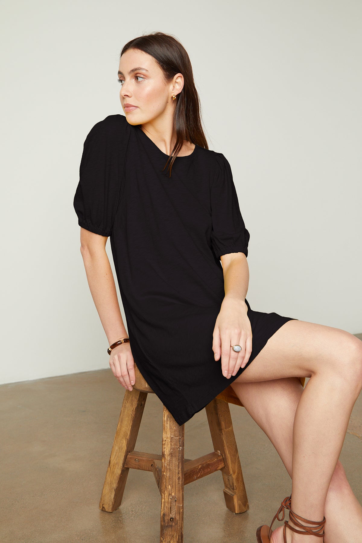   Model sitting on stool in studio wearing Meghan Dress in black with puff sleeves showing front & side 