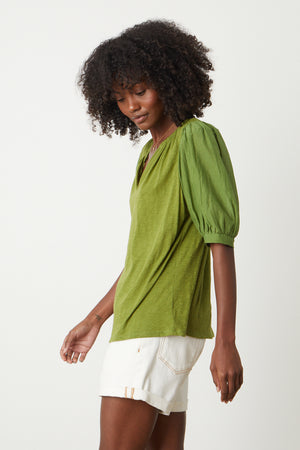 Woman sitting on stool in studio wearing Mallory Top in moss green with puff sleeves paired with white denim shorts side