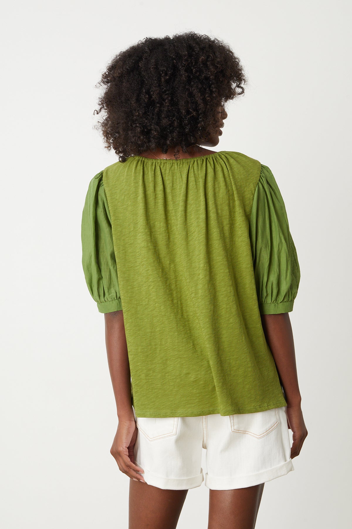   Woman sitting on stool in studio wearing Mallory Top in moss green with puff sleeves paired with white denim shorts back 